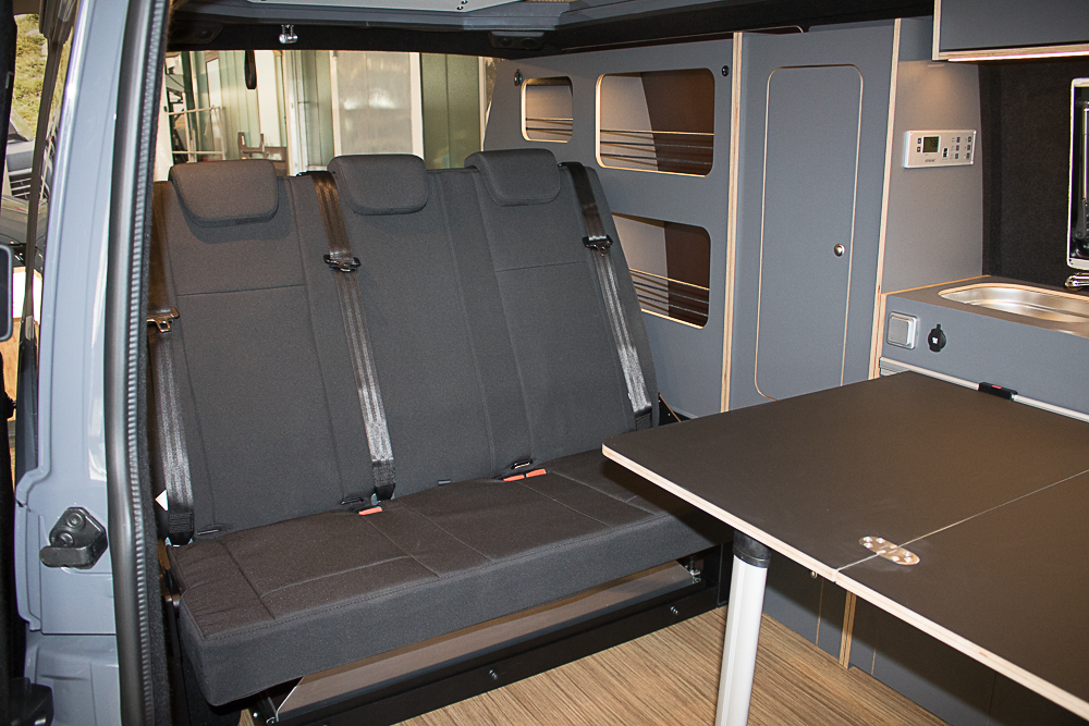 Innenraum eines Wohnmobils mit umgeklappten Sitzen, Tisch und Küchenbereich.